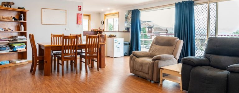 Dining and living area in a supported independent living home in Hobart, featuring a wooden dining table, comfortable seating, and large windows with views, emphasizing accessibility and community living.